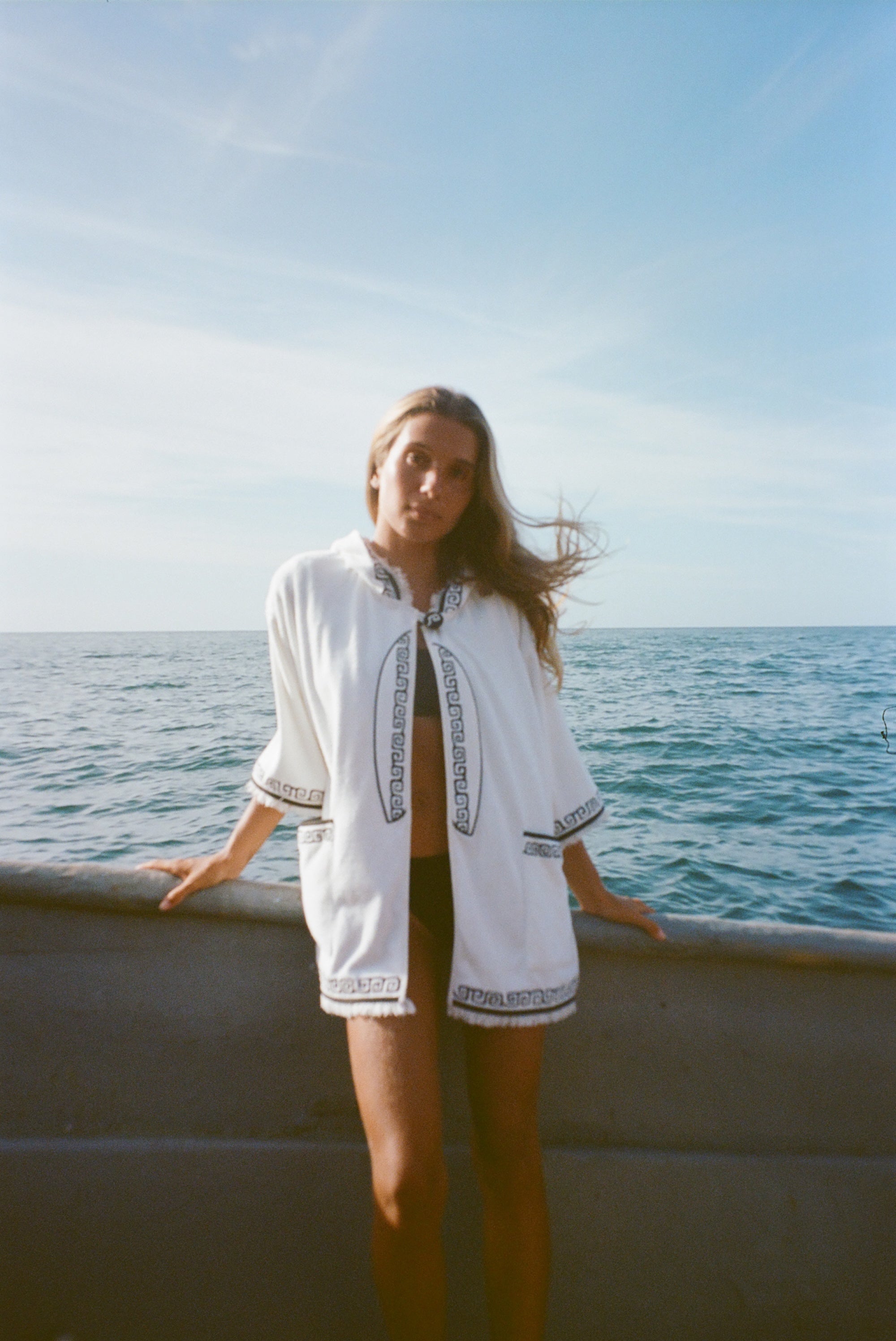 Blonde woman standing on a boat facing the camera, wearing white hooded terry cloth beach towel top with black embroidered trim, over a black bikini. 