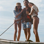 3 people standing together smiling on the edge of a boat. A male model is center, holding the rope keeping the boat at dock and wearing the 'Bisque Averse' Cabana with Tombolo floral patterned swim trunks. 