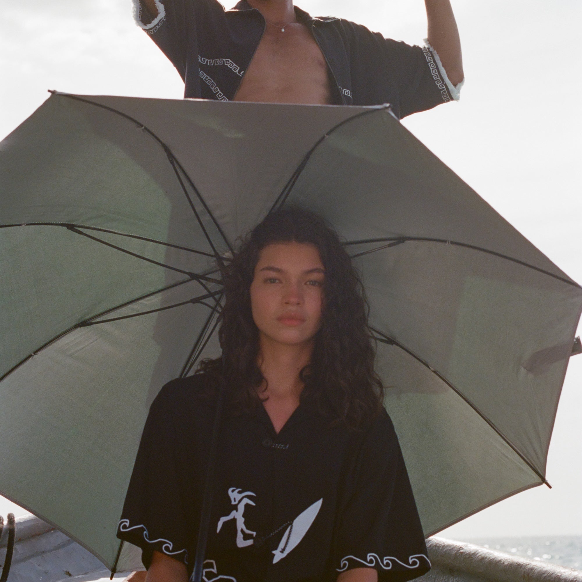Two people on a boat holding umbrellas. The woman in front wears the black shirt with white embroidered waves and a surfer design, sitting under an open umbrella. The man behind her, standing in an open black shirt with white trim, holds an umbrella above his head and wears a straw hat. 