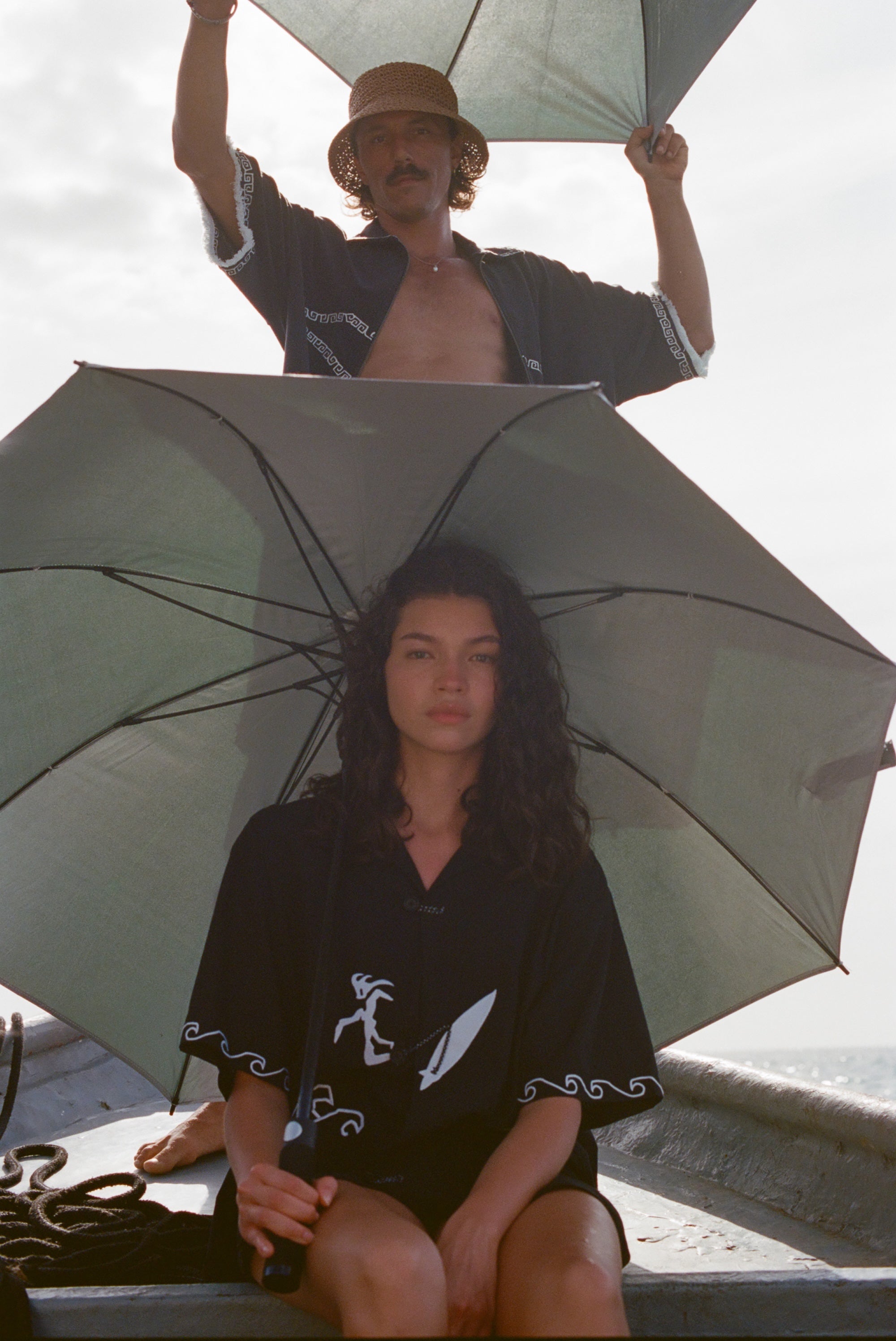 Two people on a boat holding umbrellas. The woman in front wears the black shirt with white embroidered waves and a surfer design, sitting under an open umbrella. The man behind her, standing in an open black shirt with white trim, holds an umbrella above his head and wears a straw hat. 
