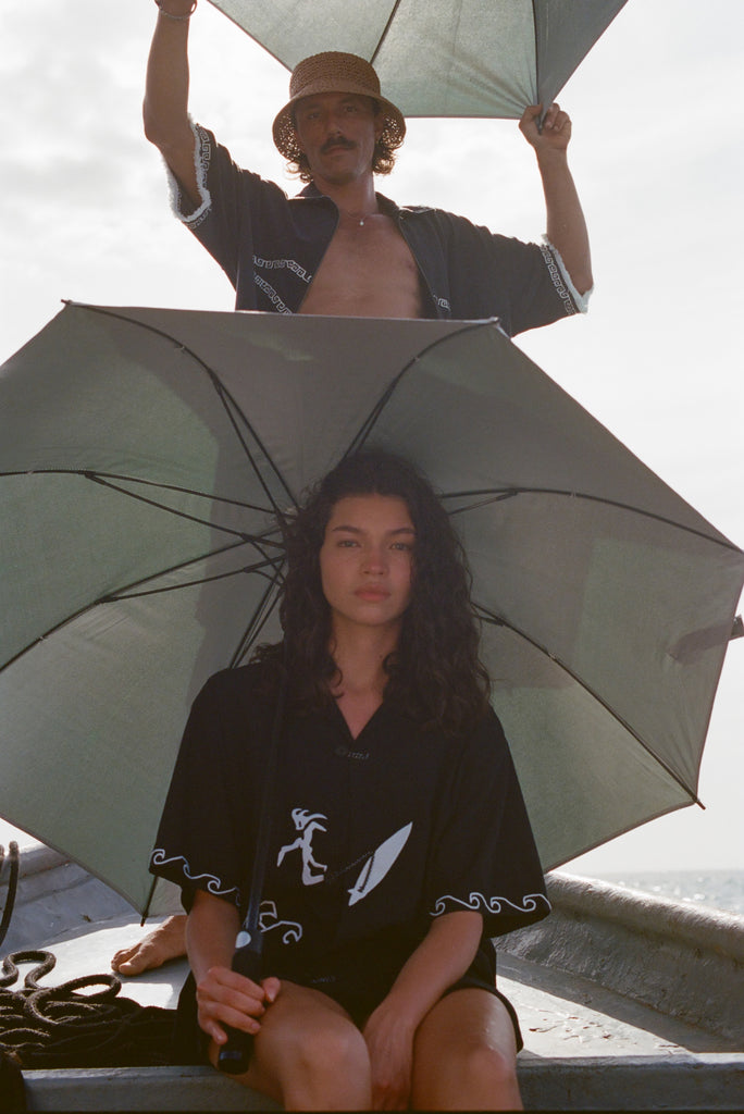 Two people on a boat holding umbrellas. The woman in front wears the black shirt with white embroidered waves and a surfer design, sitting under an open umbrella. The man behind her, standing in an open black shirt with white trim, holds an umbrella above his head and wears a straw hat. 