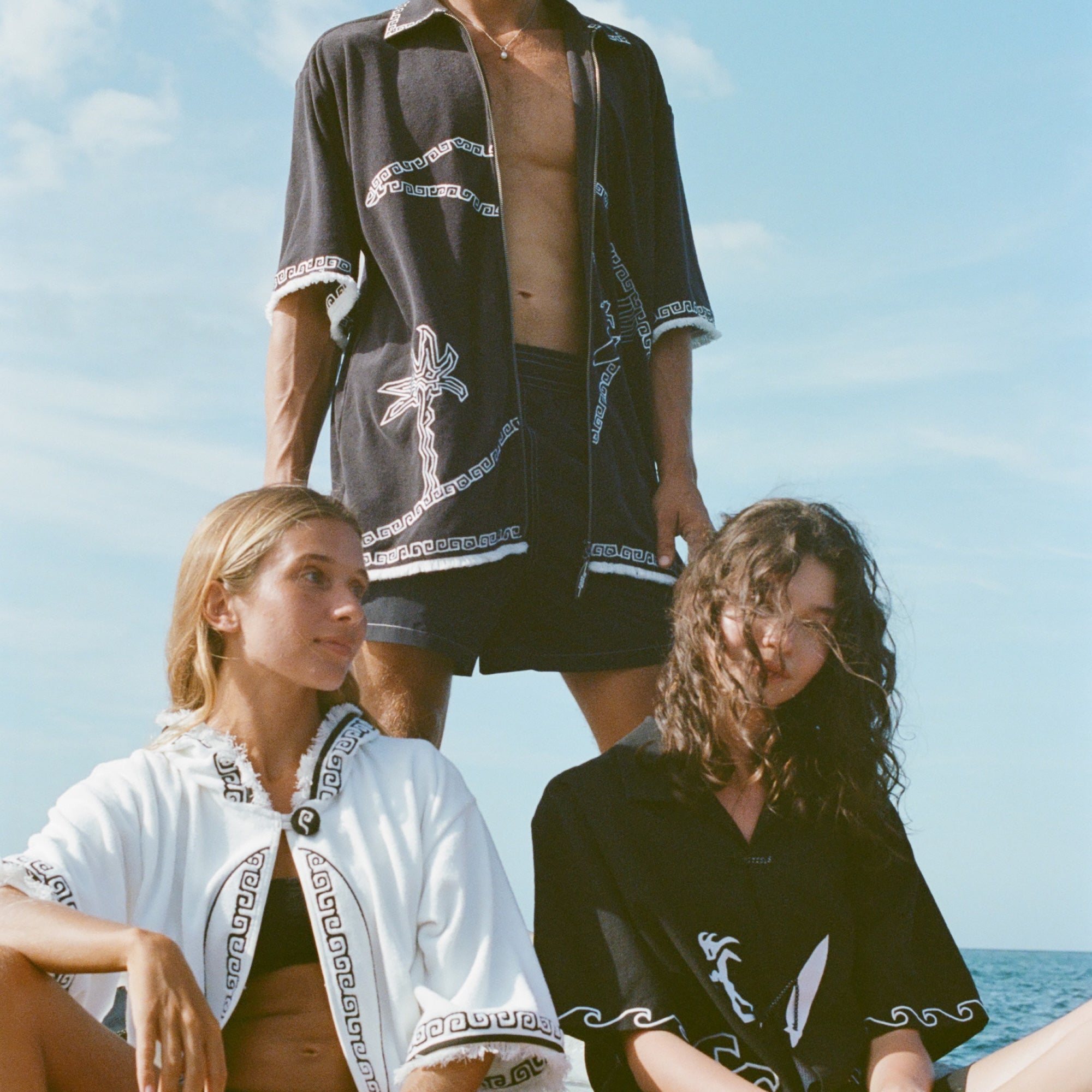 Brunette man wearing the 'Beach Break' Cabana fully unzipped. He stands behind two female models, both wearing a different product from Tombolo's Surf Collection.