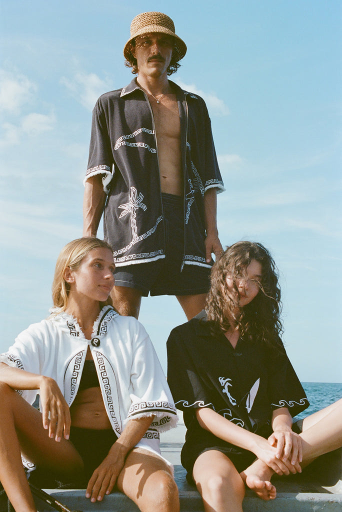 Brunette man wearing the 'Beach Break' Cabana fully unzipped. He stands behind two female models, both wearing a different product from Tombolo's Surf Collection.