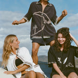 Brunette man wearing the 'Beach Break' Cabana, and a sun hat. He appears to be dancing behind two female models, one wearing the 'Surf's Up' Beach Towel Top and the other wearing the 'Unleashed' Shirt. 