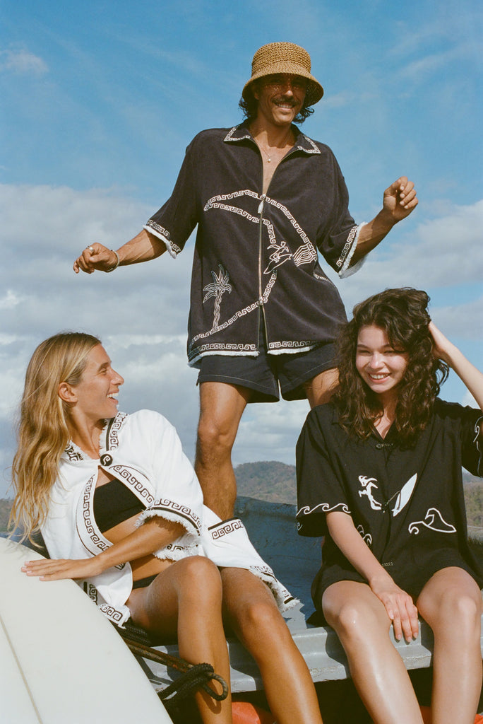 Brunette man wearing the 'Beach Break' Cabana, and a sun hat. He appears to be dancing behind two female models, one wearing the 'Surf's Up' Beach Towel Top and the other wearing the 'Unleashed' Shirt. 