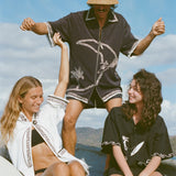 Brunette man wearing the 'Beach Break' Cabana, and a sun hat. He appears to be dancing behind two female models, both wearing a different product from Tombolo's Surf Collection.