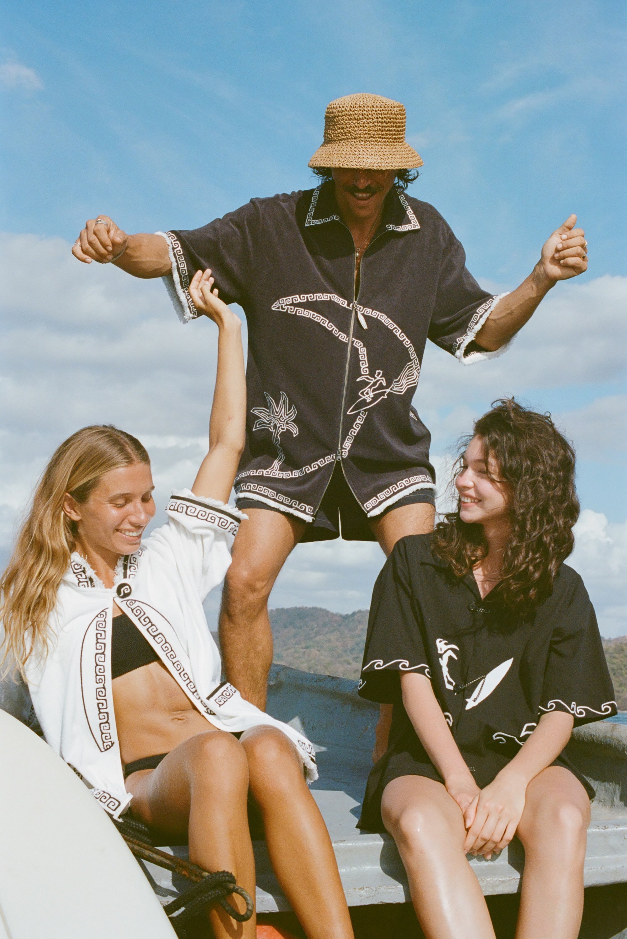 Brunette man wearing the 'Beach Break' Cabana, and a sun hat. He appears to be dancing behind two female models, both wearing a different product from Tombolo's Surf Collection.