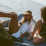 Two women on a small boat, one is in a white beach towel top open over a black bikini and straw hat, the other sitting beside her wearing the black short-sleeve terry cloth surf cabana.