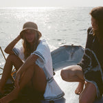 Two women on a small boat, one in a white beach towel top open over a black bikini and straw hat, the other sitting beside her wearing a black short-sleeve terry cloth surf cabana.