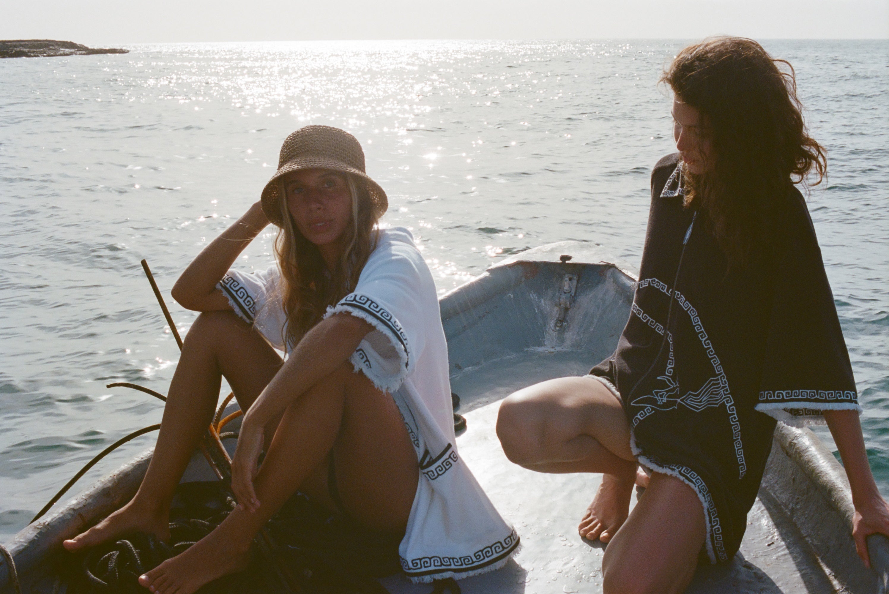 Two women on a small boat, one in a white beach towel top open over a black bikini and straw hat, the other sitting beside her wearing a black short-sleeve terry cloth surf cabana.