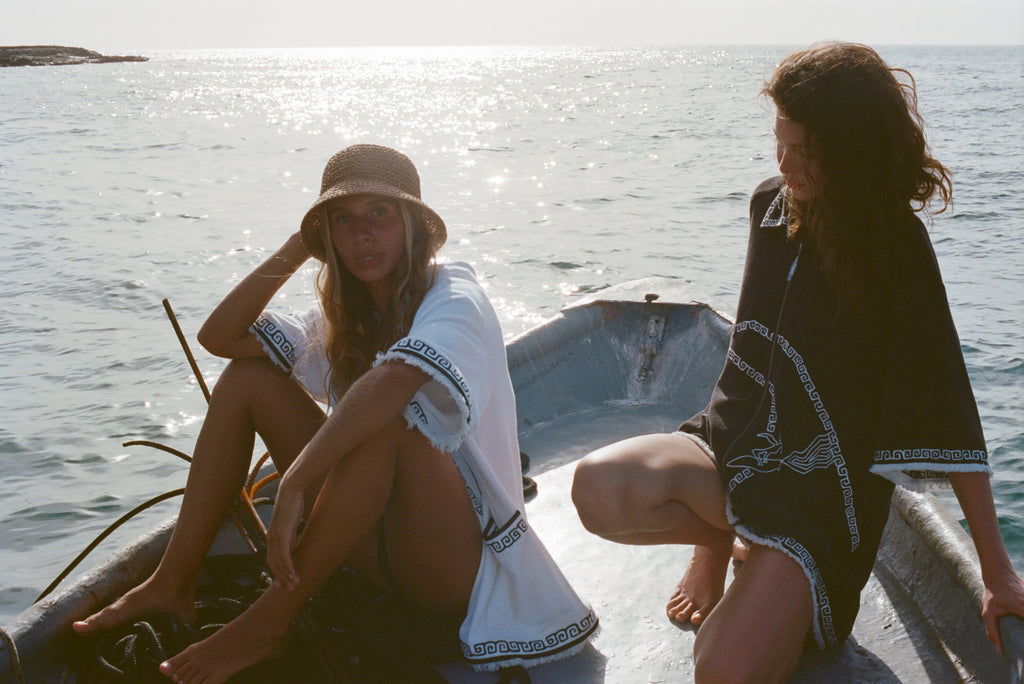 Two women on a small boat, one in a white beach towel top open over a black bikini and straw hat, the other sitting beside her wearing a black short-sleeve terry cloth surf cabana.