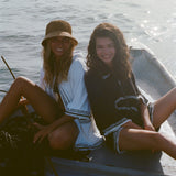 Two women sitting side by side on a boat, smiling toward the camera. One wears a white beach towel top and a straw bucket hat, the other wears a black short-sleeve terry cloth 'Beach Break' Cabana.