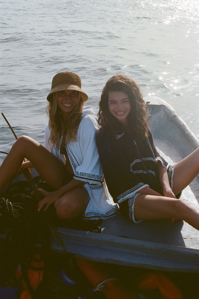 Two women sitting side by side on a boat, smiling toward the camera. One wears a white beach towel top and a straw bucket hat, the other wears a black short-sleeve terry cloth 'Beach Break' Cabana.