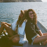 Two women on a small boat, one in a white beach towel top open over a black bikini and straw hat, the other sitting beside her wearing a black short-sleeve terry cloth surf cabana. Both are smiling at the camera.