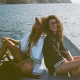 Two women on a small boat, one in a white beach towel top open over a black bikini and straw hat, the other sitting beside her wearing a black short-sleeve terry cloth surf cabana. Both are smiling at the camera.