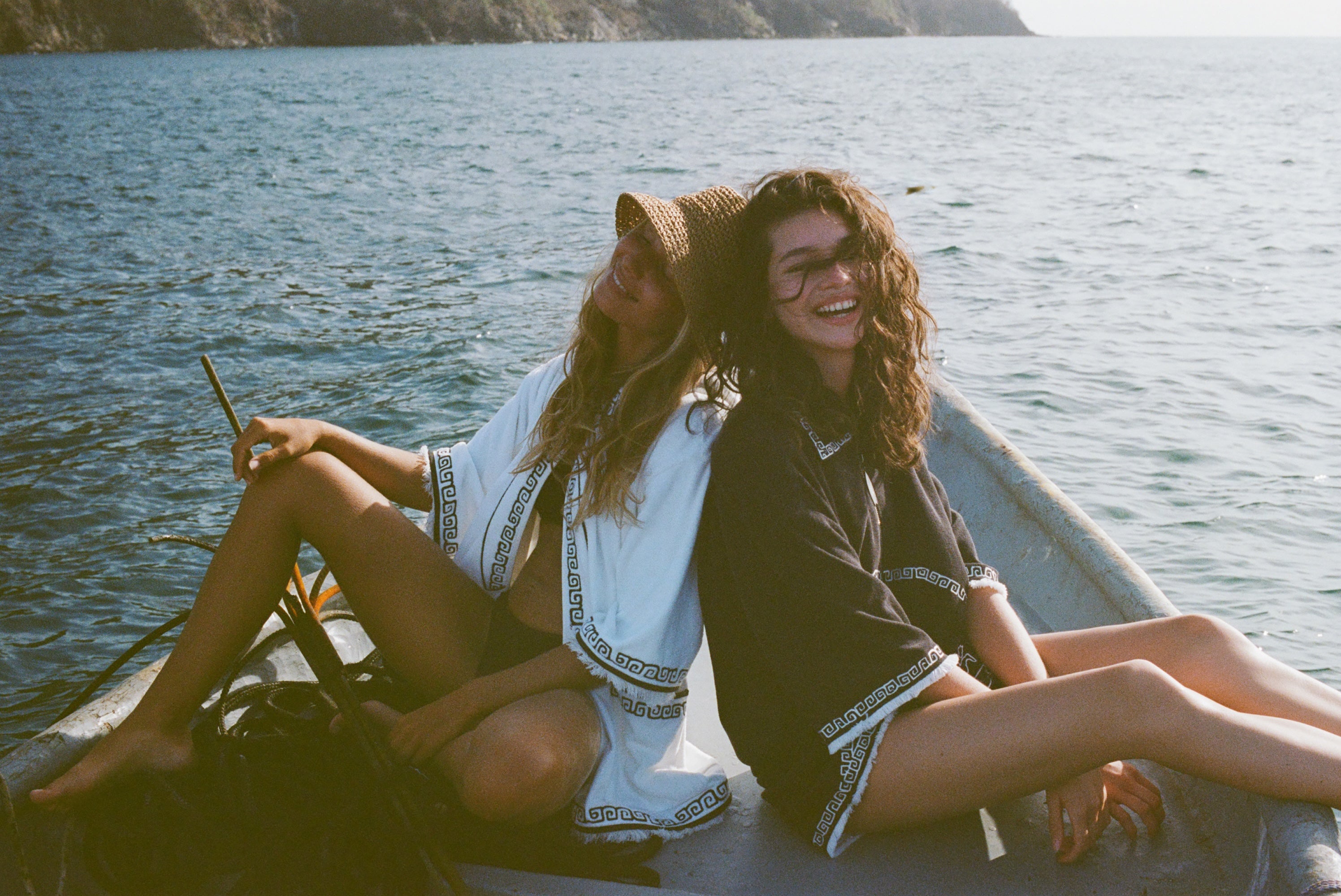 Two women on a small boat, one in a white beach towel top open over a black bikini and straw hat, the other sitting beside her wearing a black short-sleeve terry cloth surf cabana. Both are smiling at the camera.