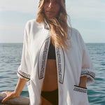 Blonde woman standing on a boat, wearing white hooded terry cloth beach towel top with black embroidered trim, over a black bikini.