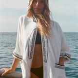 Blonde woman standing on a boat, wearing white hooded terry cloth beach towel top with black embroidered trim, over a black bikini.