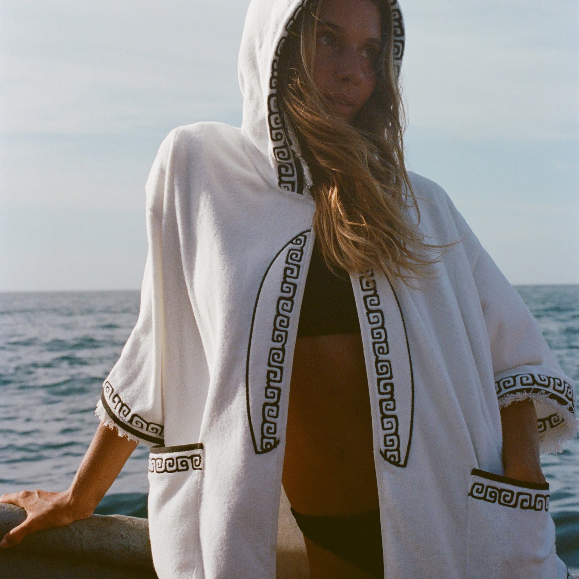 Blonde woman standing on a boat, wearing white hooded terry cloth beach towel top with black embroidered trim, over a black bikini. She is looking off to the side. 
