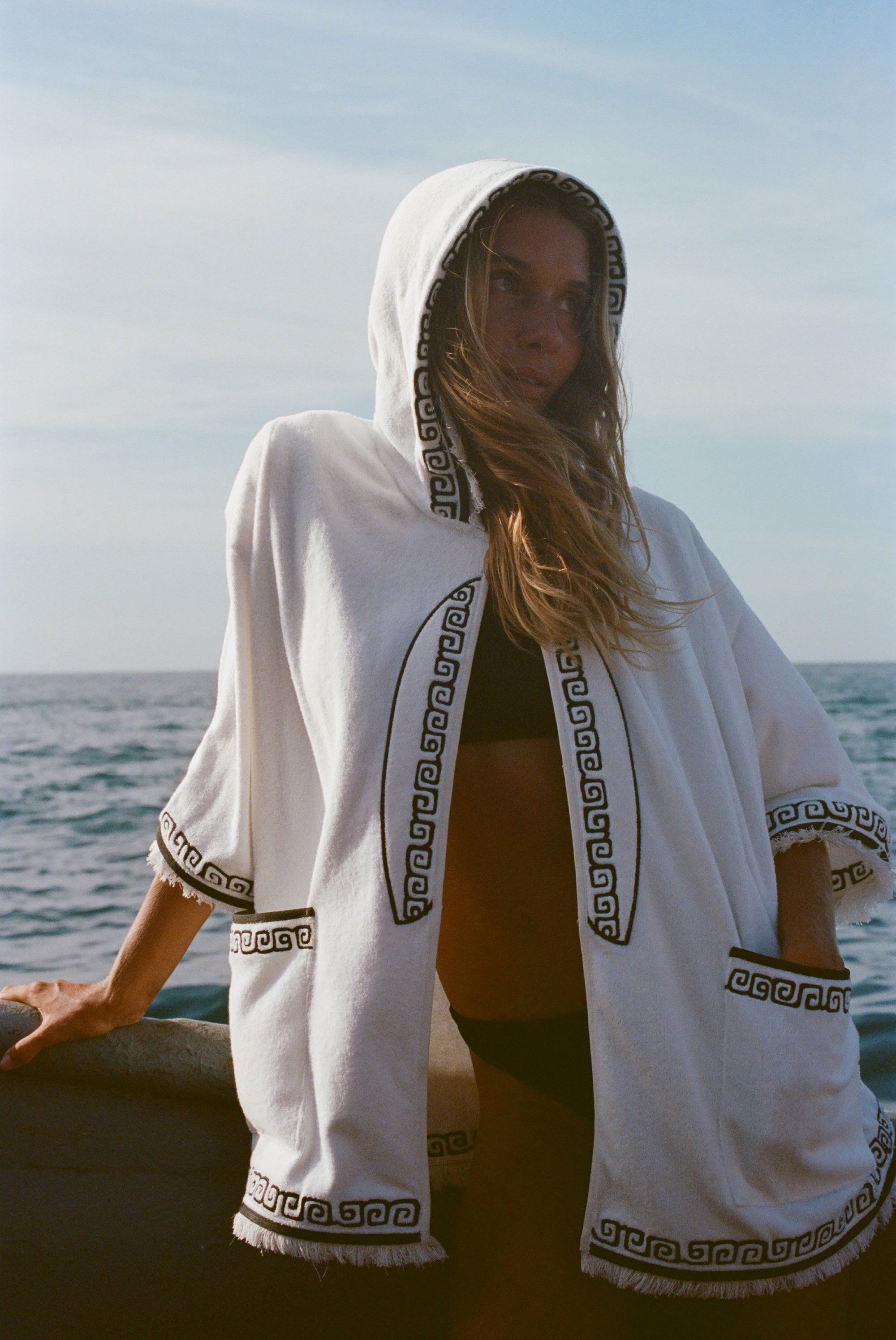 Blonde woman standing on a boat, wearing white hooded terry cloth beach towel top with black embroidered trim, over a black bikini. She is looking off to the side. 