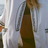 Detail image of woman wearing white surf beach towel top over a black bikini. This image shows more details of the black embroidered surfboard featured on the front.