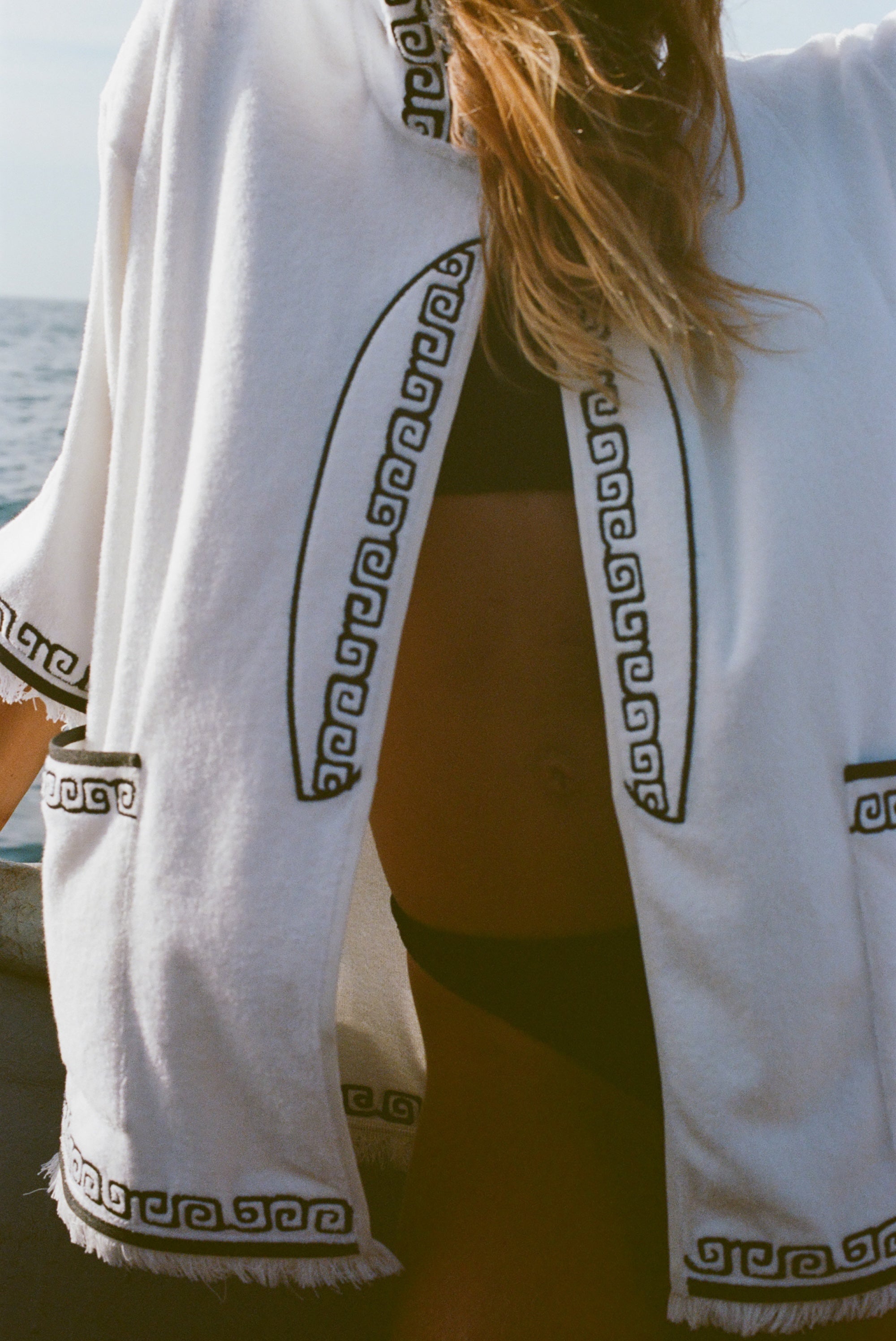 Detail image of woman wearing white surf beach towel top over a black bikini. This image shows more details of the black embroidered surfboard featured on the front.