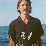 Man standing on a boat wearing a black short-sleeve shirt with white embroidered waves and a surfer with a board attached by an ankle leash.