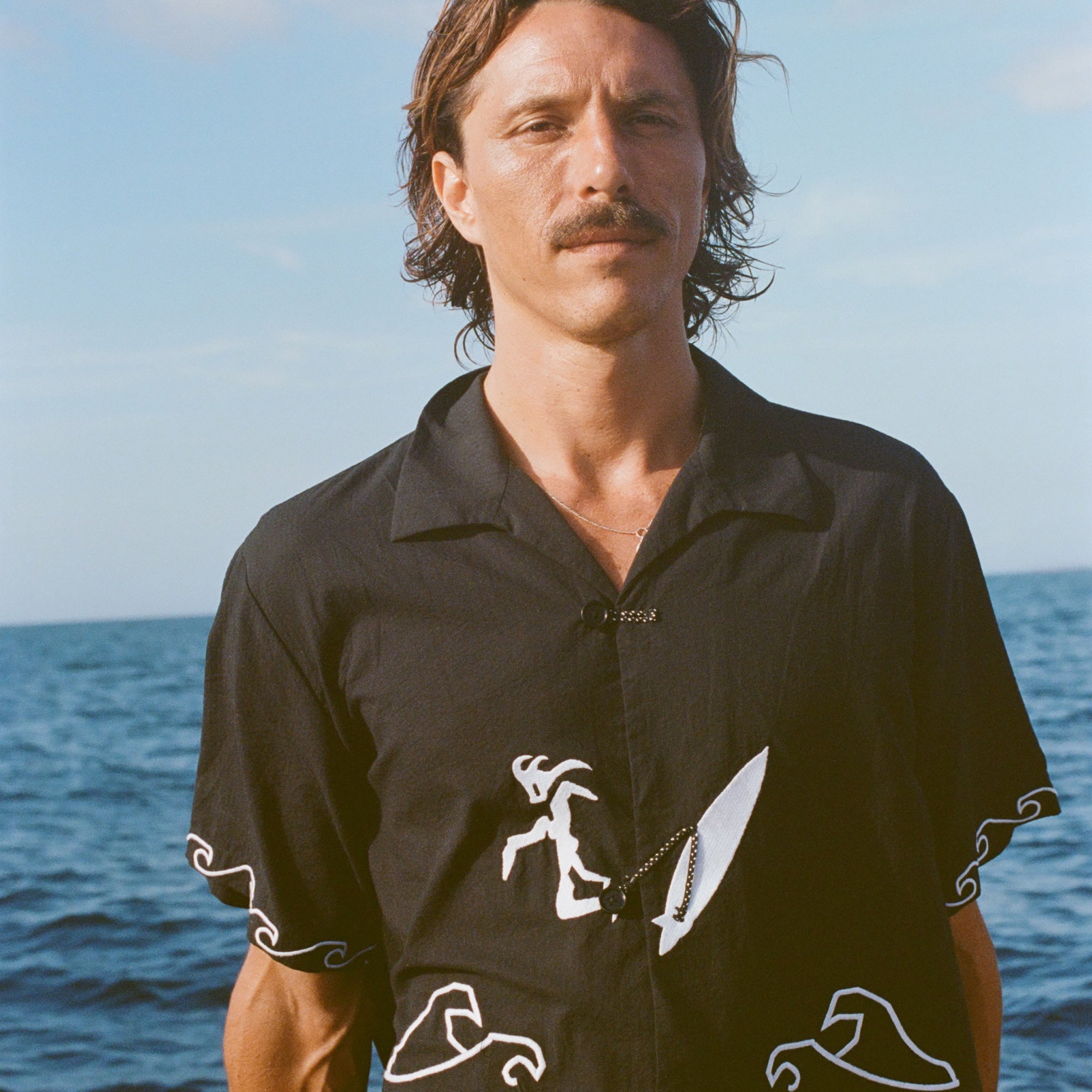 Man standing on a boat wearing a black short-sleeve shirt with white embroidered waves and a surfer with a board attached by an ankle leash.