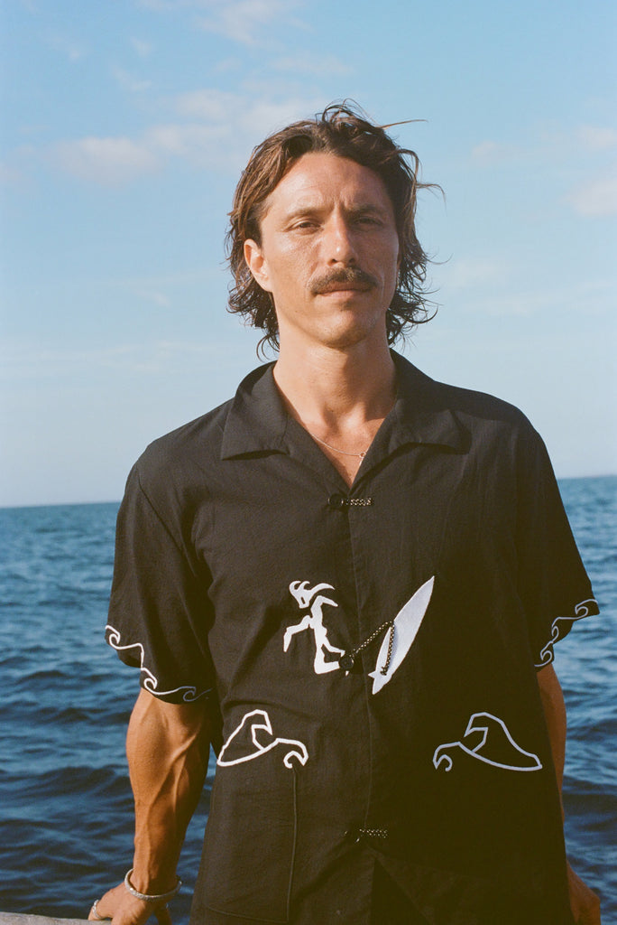 Man standing on a boat wearing a black short-sleeve shirt with white embroidered waves and a surfer with a board attached by an ankle leash.