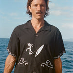 Man standing on a boat wearing the black short-sleeve shirt with white embroidered surfer and wave designs. The shirt features knotted loop closures.