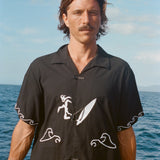 Man standing on a boat wearing the black short-sleeve shirt with white embroidered surfer and wave designs. The shirt features knotted loop closures.
