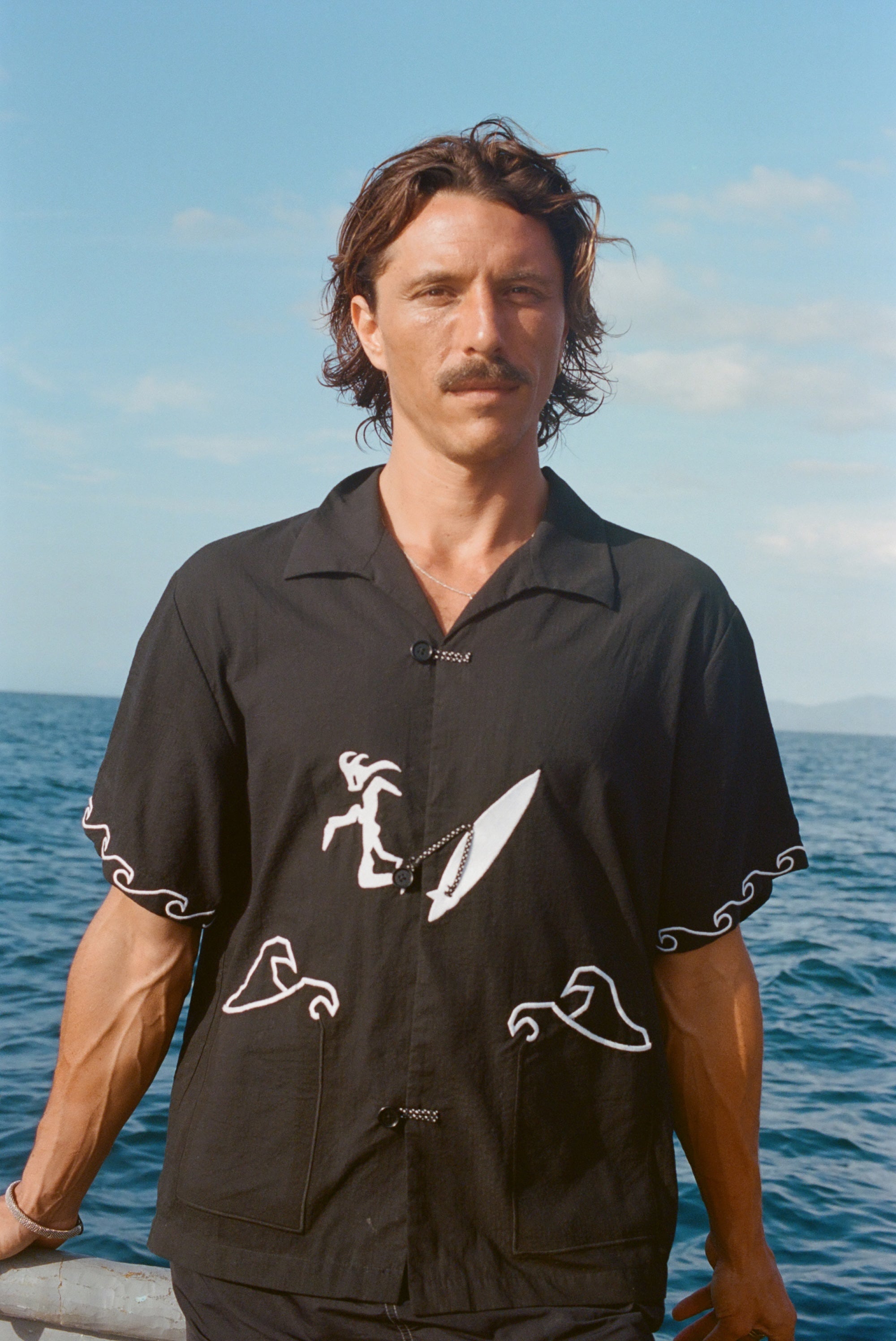 Man standing on a boat wearing the black short-sleeve shirt with white embroidered surfer and wave designs. The shirt features knotted loop closures.