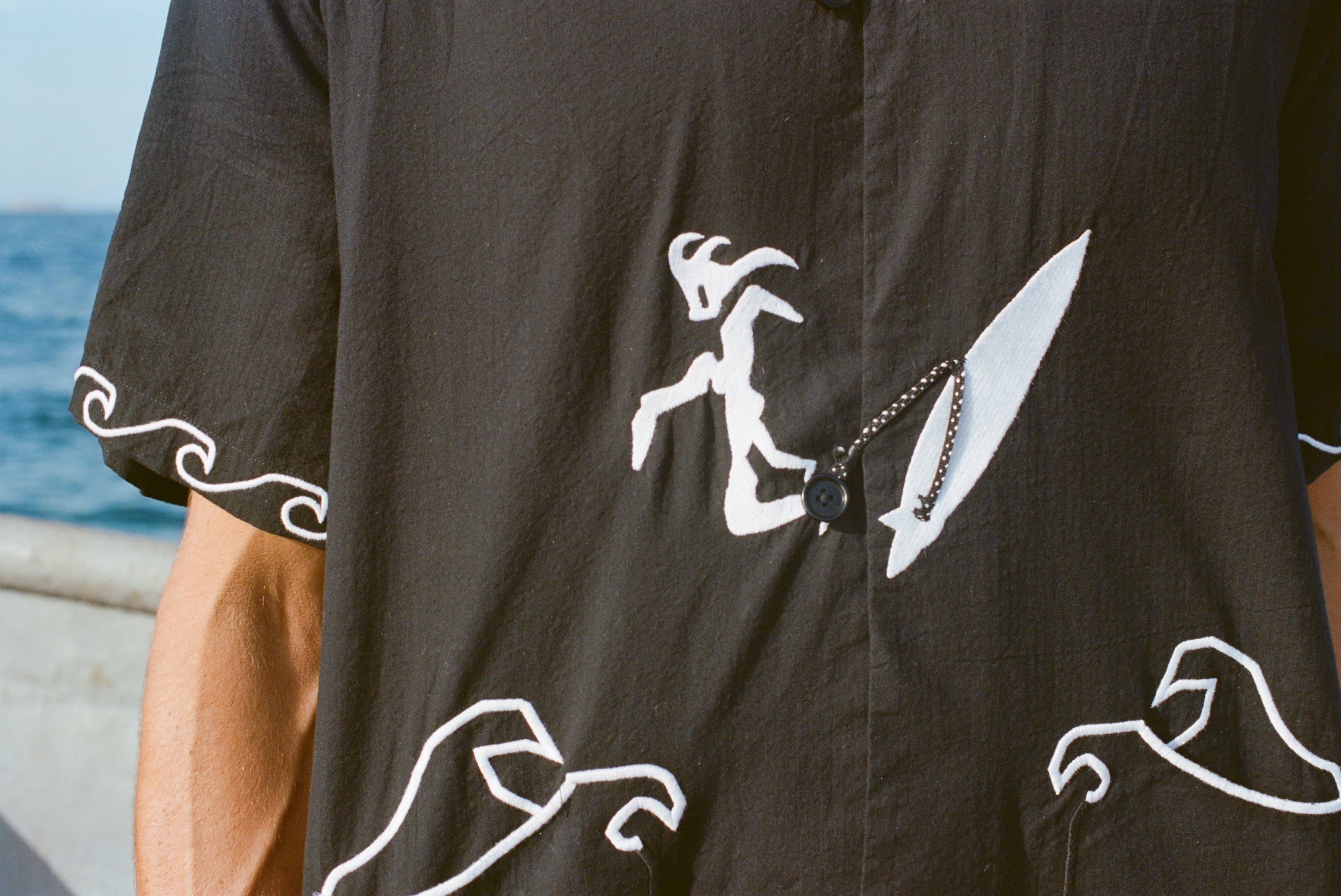 Close-up image of the embroidered surfer, attached to his board with an ankle leash. The ankle leash appears as a small loop to button the shirt.
