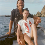 A man and woman sit together at the edge of a small boat on the water. The man, seated behind, wears a black short-sleeve terry cloth shirt with cream-colored embroidery featuring a stylized figure and agave plant ('Agua Magica' Cabana). The woman, leaning back against him, wears a sleeveless white terry cloth top with a bold red lobster design on each side of the chest and red trim ('Better Half' Hand Towel Top) 