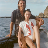 A man and woman sit together at the edge of a small boat on the water. The man, seated behind, wears a black short-sleeve terry cloth shirt with cream-colored embroidery featuring a stylized figure and agave plant ('Agua Magica' Cabana). The woman, leaning back against him, wears a sleeveless white terry cloth top with a bold red lobster design on each side of the chest and red trim ('Better Half' Hand Towel Top) 