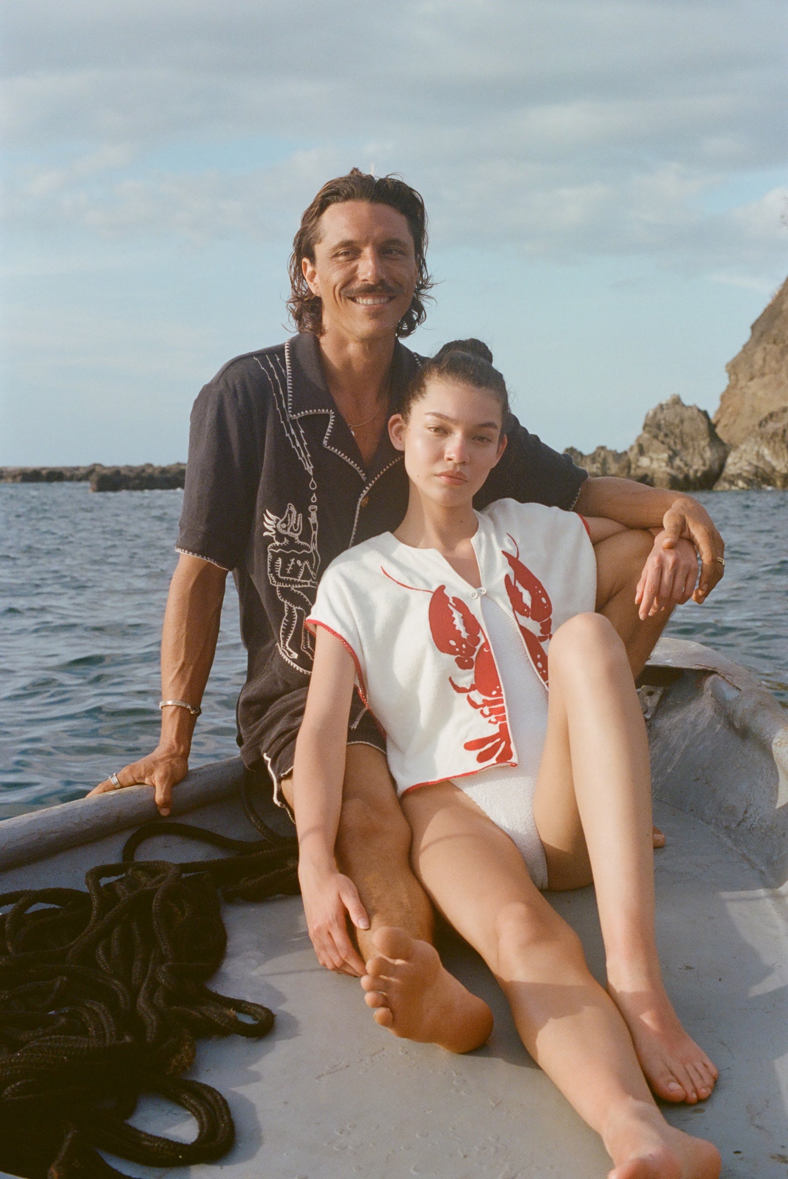 A man and woman sit together at the edge of a small boat on the water. The man, seated behind, wears a black short-sleeve terry cloth shirt with cream-colored embroidery featuring a stylized figure and agave plant ('Agua Magica' Cabana). The woman, leaning back against him, wears a sleeveless white terry cloth top with a bold red lobster design on each side of the chest and red trim ('Better Half' Hand Towel Top) 