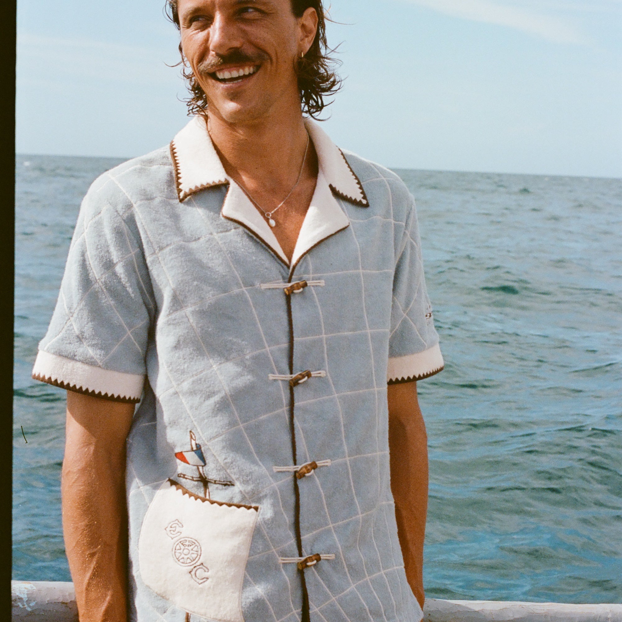 Male model smiling on boat, wearing 'The Explorers Club' Expedition Cabana and swim trunks.