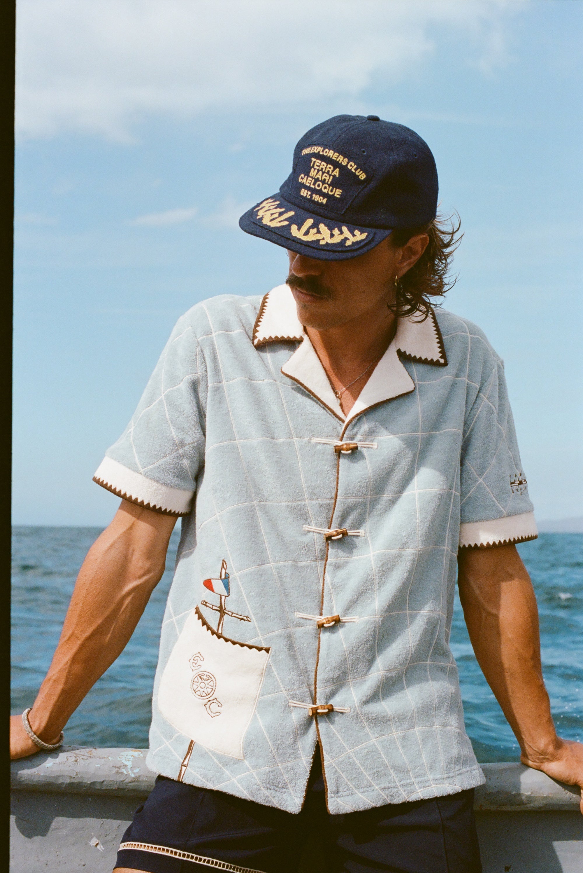 Male model wearing 'The Explorers Club' 6-Panel Souvenir Cap (Wool) with 'The Explorers Club' Expedition Cabana, leaning on the side of a boat in the water.