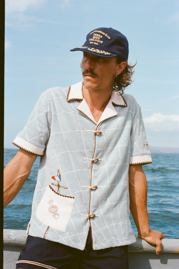 Male model wearing 'The Explorers Club' 6-Panel Souvenir Cap (Wool) with 'The Explorers Club' Expedition Cabana, leaning on the side of a boat in the water.