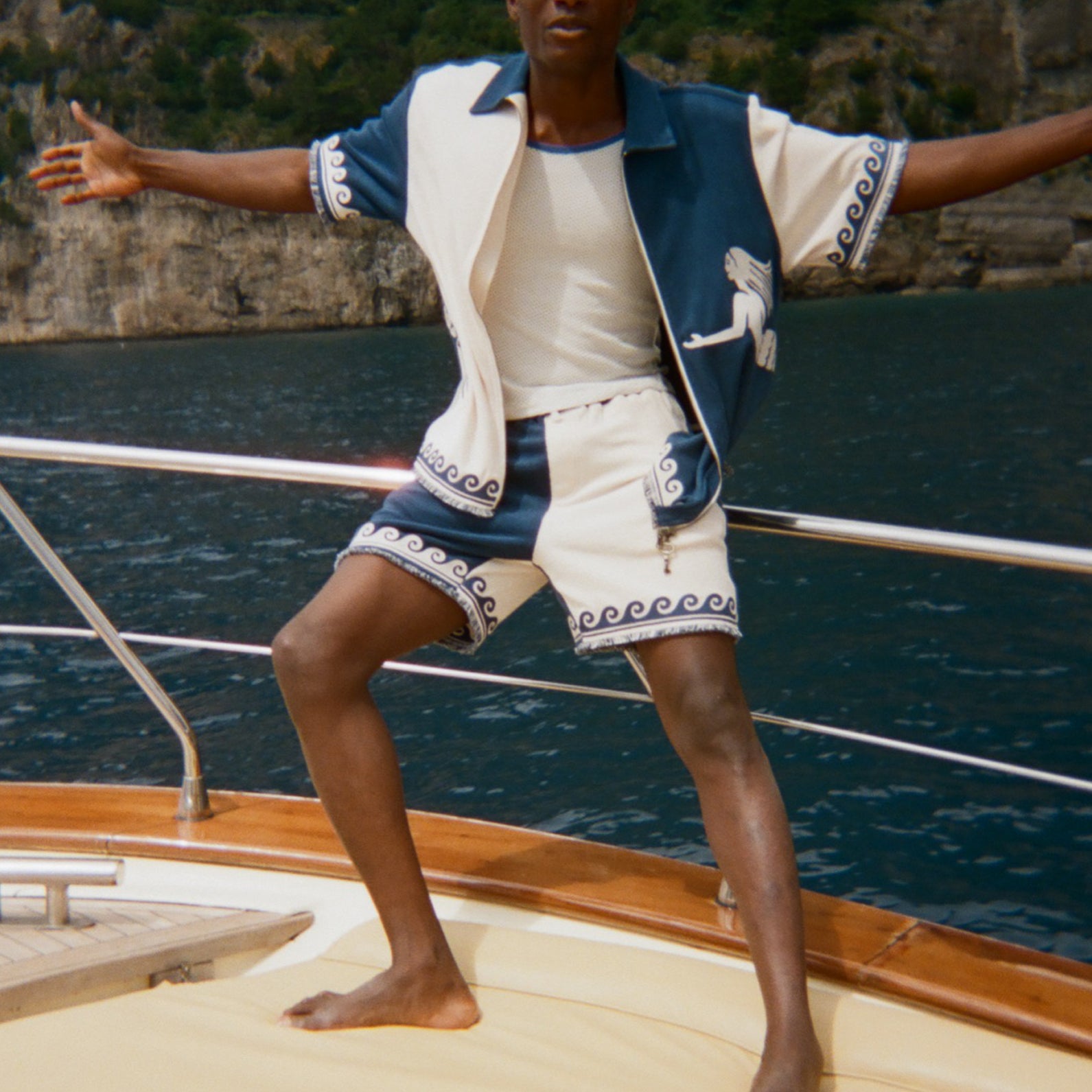 Person standing on a boat with arms outstretched, wearing a blue and white Late Checkout Cabana set. 