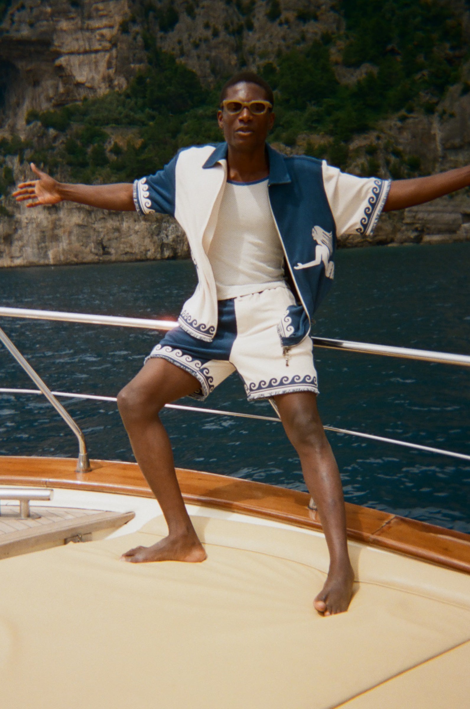 Person standing on a boat with arms outstretched, wearing a blue and white Late Checkout Cabana set. 