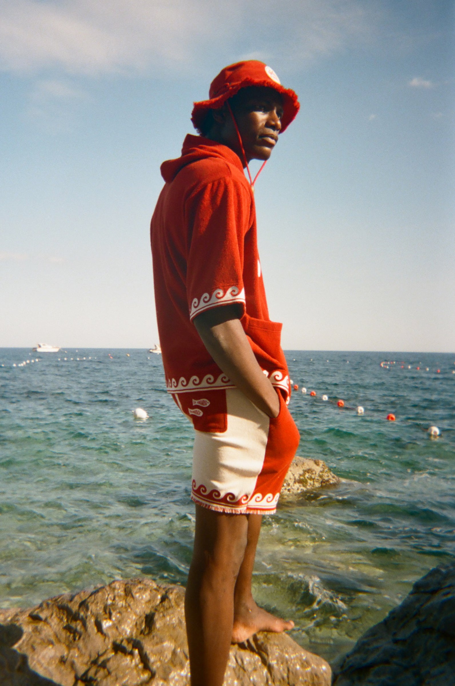 Person wearing a red Le Sirenuse shirt and hat standing on a rock in front of the ocean. 
