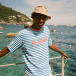 Person wearing a Le Sirenuse striped shirt and hat standing on a dock with a scenic ocean view.