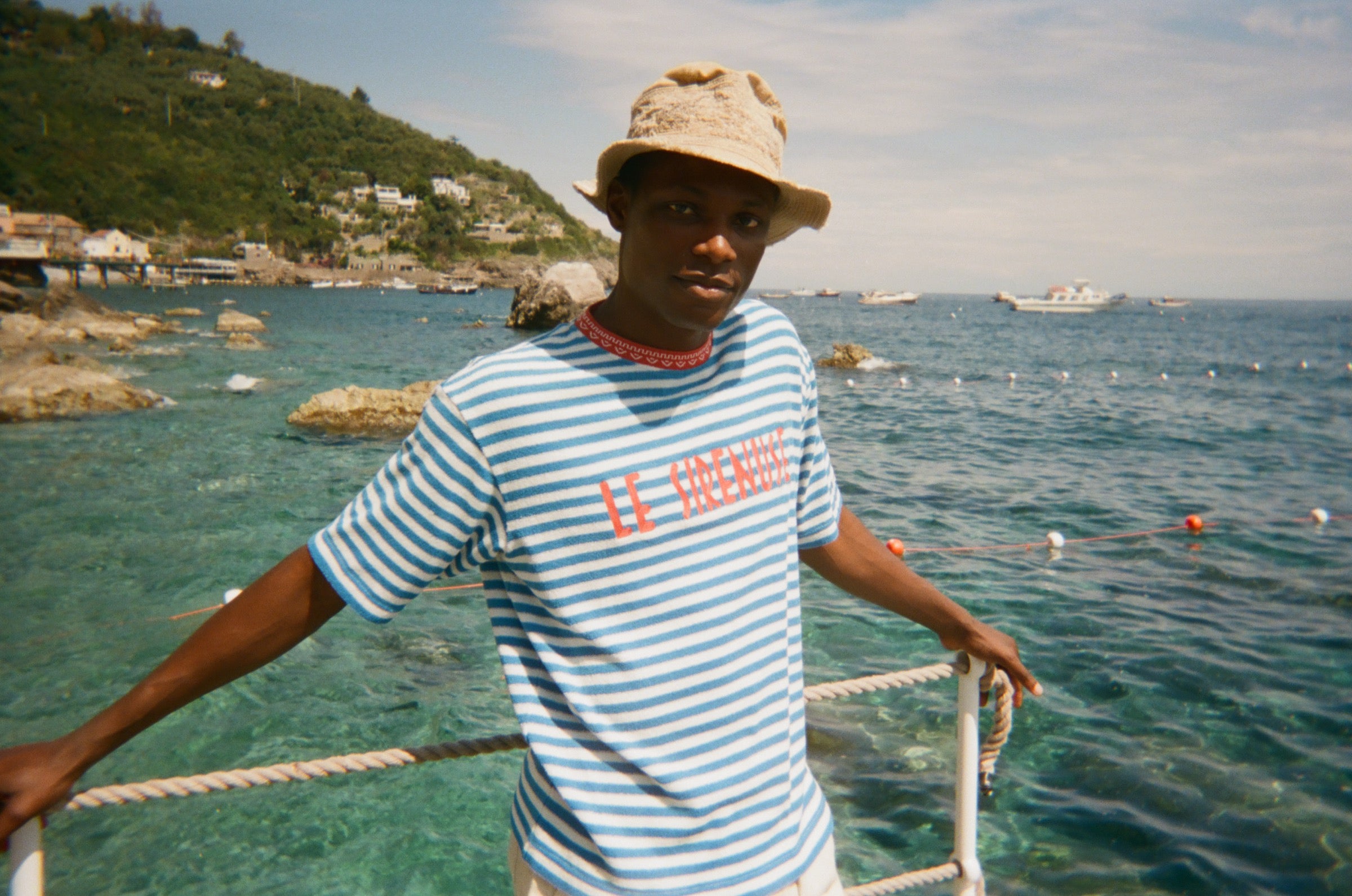 Person wearing a Le Sirenuse striped shirt and hat standing on a dock with a scenic ocean view.