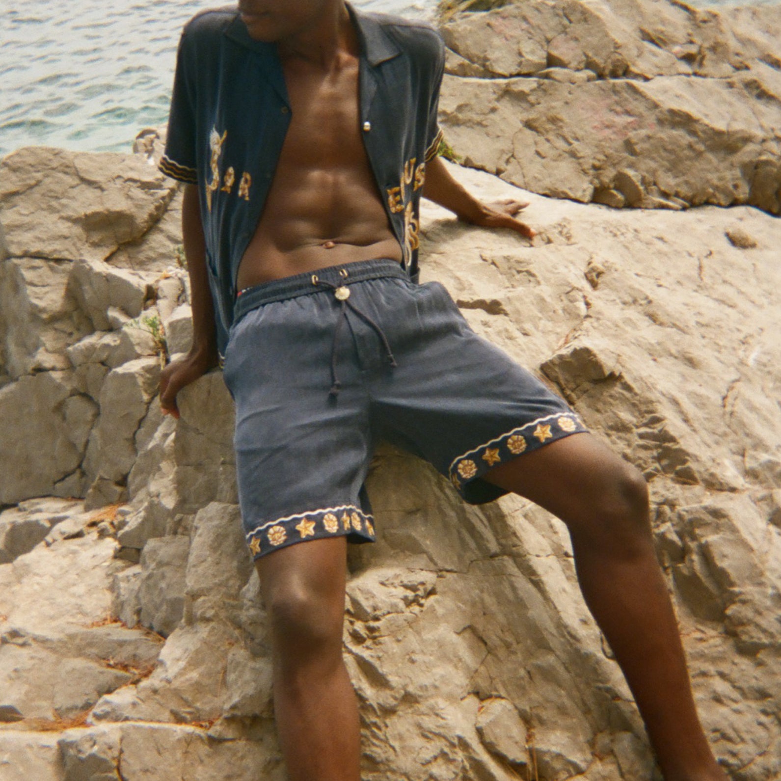 Man sitting on rocks by the water wearing a patterned shirt and shorts.