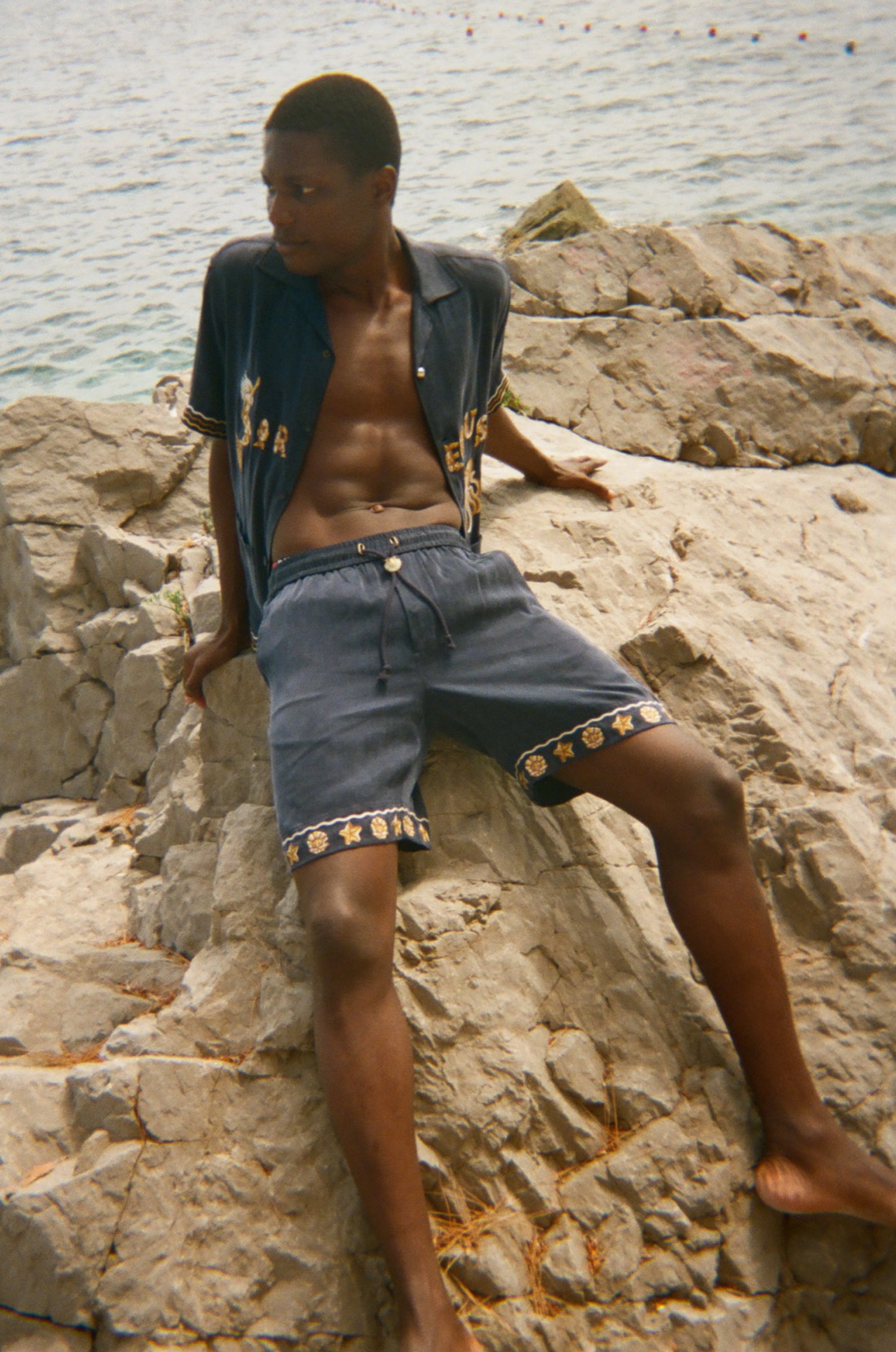 Man sitting on rocks by the water wearing a patterned shirt and shorts.