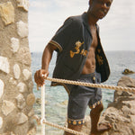 Man standing on a stone ledge by the ocean wearing a dark Le Sirenuse shirt and matching shorts. 