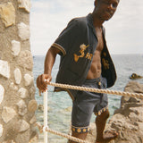 Man standing on a stone ledge by the ocean wearing a dark Le Sirenuse shirt and matching shorts. 