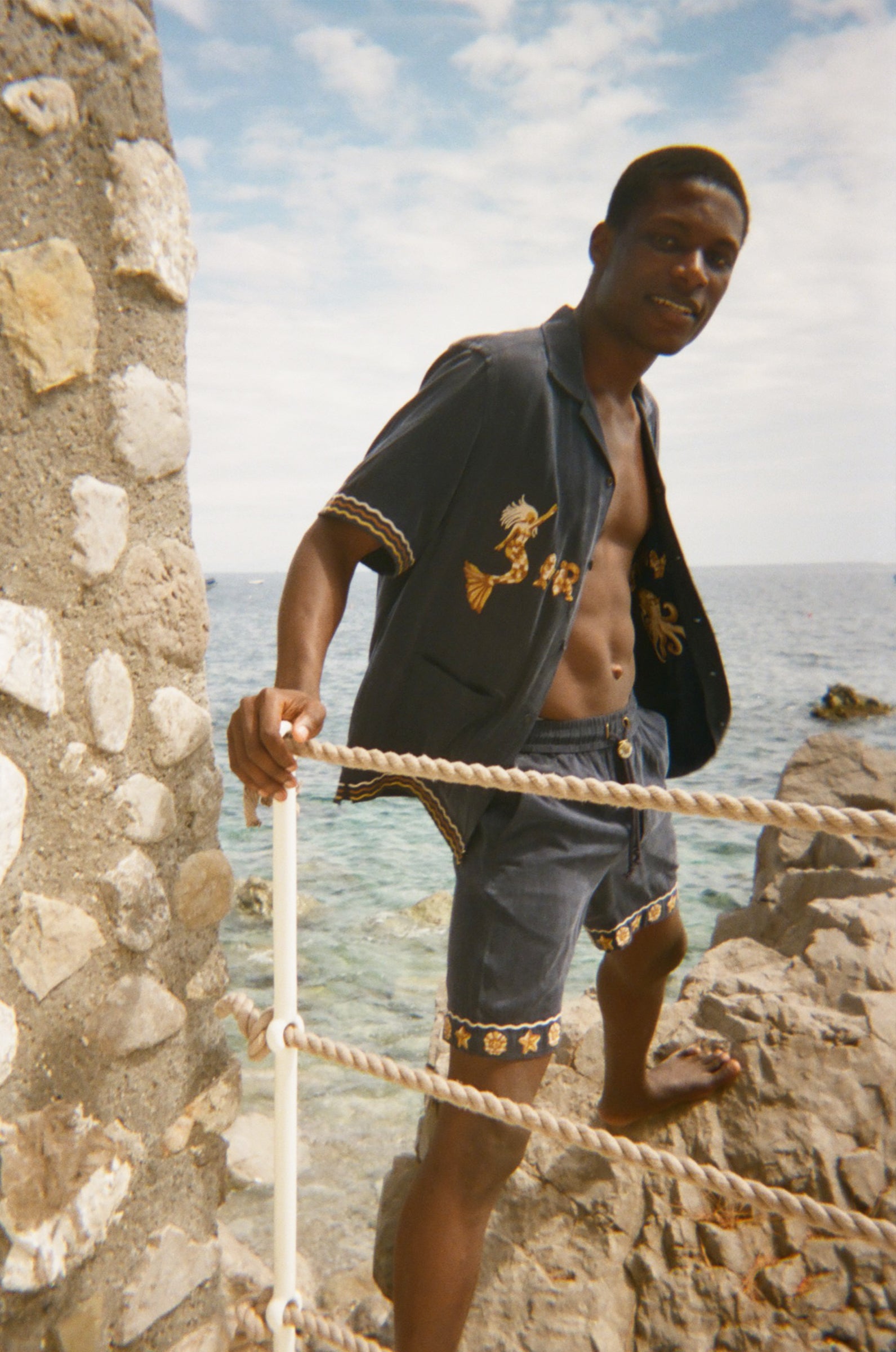 Man standing on a stone ledge by the ocean wearing a dark Le Sirenuse shirt and matching shorts. 