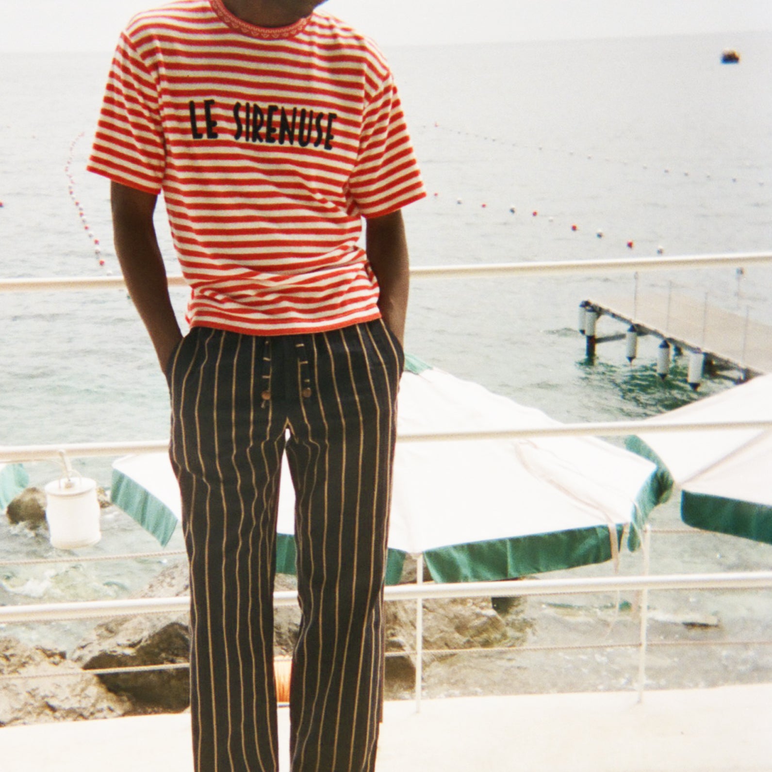 Person wearing a red and white striped shirt with 'Le Sirenuse' embroidered, and black and white striped pants standing on a deck.